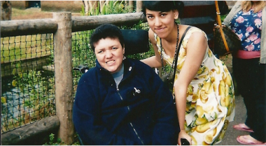 Two people, one in a wheelchair and the other standing, are smiling next to each other.