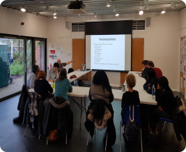 A gathering of individuals seated in a classroom-like setting, attentively watching a presentation on a projector screen.